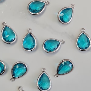 Può includere: Ciondoli in vetro turchese a forma di goccia con finiture in metallo color argento. I ciondoli sono disposti su una superficie colorata chiara. Ogni ciondolo ha un piccolo anello nella parte superiore per il fissaggio.