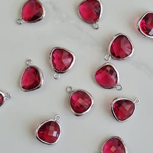 Könnte beinhalten: Eine Sammlung facettierter, herzförmiger, rubinroter Anhänger mit silberfarbenem Metallbesatz und kleinen Schlaufen zum Befestigen an Schmuck. Die Anhänger sind auf einer weißen Oberfläche verstreut.