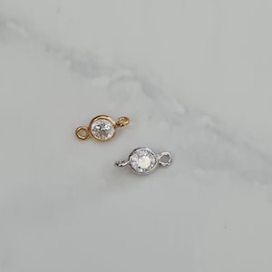 Op de afbeelding: Twee sieradenverbinders met een heldere ronde strass in het midden. De ene connector is goud en de andere is zilver.