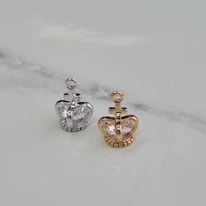May include: Two small crown charms, one silver and one gold, with a clear rhinestone in the centre. Both charms have a loop at the top for attaching to a necklace or bracelet.