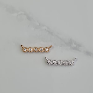 May include: Two curved jewellery pieces, one gold-toned and one silver-toned, each with five clear, round gemstones. Small loops are on each end. The background is a light, neutral colour.