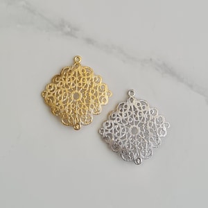May include: Two filigree metal charms, one gold and one silver, in the shape of a square with a decorative floral design.