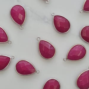 Op de afbeelding: Close-up van verschillende druppelvormige magenta edelsteenhangers met zilverkleurige metalen rand. Elke hanger heeft een facetgeslepen oppervlak en een kleine lus aan de bovenkant.