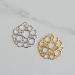 Peut inclure: Deux pendentifs décoratifs en métal. L'un est argenté, l'autre doré. Chacun présente un ensemble de cercles interconnectés de tailles variables, créant un motif en forme de bulles. Les pendentifs sont présentés sur une surface en marbre blanc.