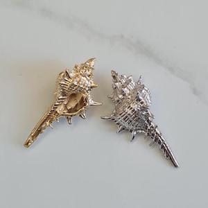 May include: Two seashell-shaped ornaments. One is gold-colored, and the other is silver-colored. Both have a detailed, textured surface with pointed edges. The ornaments are likely decorative items or jewelry components.