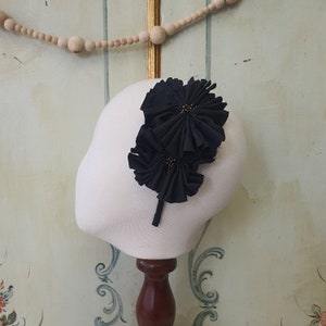 Puede incluir: Diadema de flores de tela negra con detalles dorados. La diadema se muestra en una cabeza de maniquí blanca.