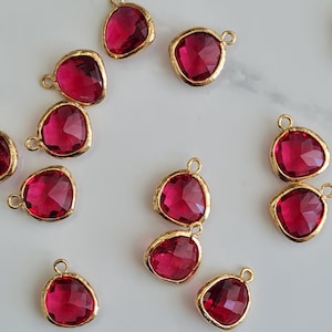 Peut inclure: Une collection de pendentifs en pierres précieuses rouge rubis avec des bordures en métal doré. Chaque pendentif a une forme de goutte d'eau facettée et une petite boucle pour l'attacher à des bijoux. Les pendentifs sont dispersés sur une surface blanche.