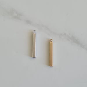 Peut inclure: Deux pendentifs en forme de barre métallique, l'un argenté et l'autre doré, chacun avec une petite boucle en haut. Les pendentifs sont rectangulaires et présentent un design simple et minimaliste. Ils sont présentés sur une surface en marbre gris clair.