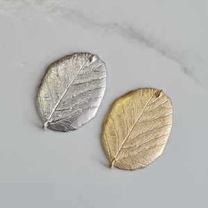 May include: Two leaf-shaped pendants, one silver and one gold. Each has detailed veining and a textured surface. The pendants are oval and suitable for jewellery making or crafts. Measurements are in centimetres.