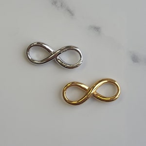 Peut inclure: Deux breloques en forme de symbole infini, l'une argentée et l'autre dorée. Les breloques sont sur un fond blanc.