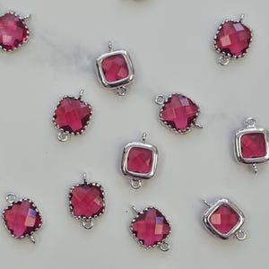 Pode incluir: Uma coleção de pingentes de pedras preciosas vermelho rubi com engastes de metal prateado. Os pingentes têm uma variedade de formas, incluindo designs quadrados e facetados. Cada pingente tem um pequeno laço para fixar.