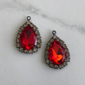 May include: Two teardrop-shaped red crystal pendants with a halo of small, light brown crystals. The pendants have a dark metal setting and a loop at the top for hanging. The crystals are faceted, catching the light. Jewellery components.
