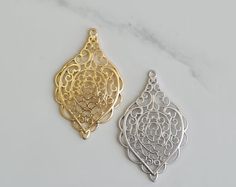 May include: Two filigree pendants, one gold-toned and one silver-toned, lie side-by-side. Each pendant has an ornate, openwork design with a teardrop shape and a small loop at the top for hanging. The pendants are likely for jewelry making.