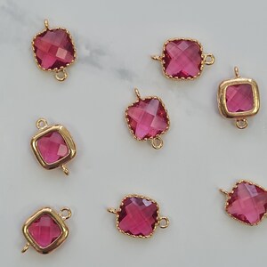 May include: A collection of gold-coloured square charms, each featuring a faceted magenta gemstone. These charms have loops at the top and bottom for attaching to jewellery. They are arranged on a light-coloured surface.