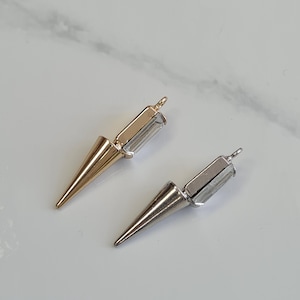 Puede incluir: Dos colgantes metálicos con forma de punta, uno dorado y otro plateado, con un acento de cristal rectangular. Los colgantes tienen un lazo en la parte superior para sujetarlos a la joyería. El fondo es una superficie clara, similar al mármol.