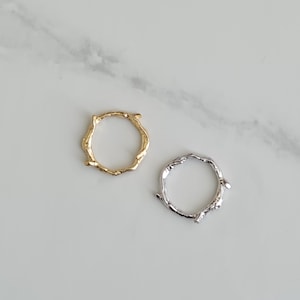 May include: Two rings, one gold and one silver, are shown on a white marble surface. The rings have a twig-like design with irregular, textured surfaces. The gold ring is on the left, and the silver ring is on the right.