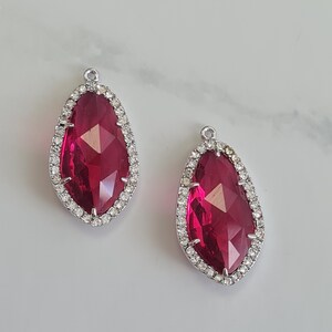 May include: Two teardrop-shaped pendants. Each features a faceted, deep red centre stone, framed by a silver-coloured setting with small, clear crystals. A small loop at the top allows attachment to a chain or other jewellery component.