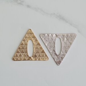 以下が含まれることがあります： 金色の三角形と銀色の三角形の2つの金属製ペンダント。中央に楕円形の切り抜きがあり、どちらも点状のテクスチャパターンが施されています。ジュエリー作りやクラフトに最適です。