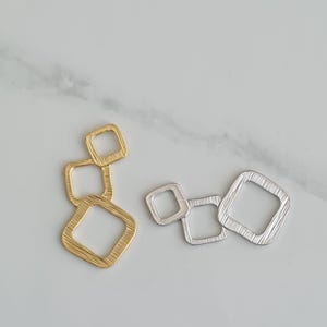 Puede incluir: Dos juegos de dijes metálicos geométricos. Un juego es dorado y presenta tres cuadrados conectados. El otro juego es plateado y presenta tres cuadrados conectados.