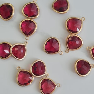 May include: Close-up of several heart-shaped ruby red gemstone pendants with gold-coloured bezels. Each pendant has a small loop at the top for attaching to a chain or other jewellery component. The gemstones are faceted.
