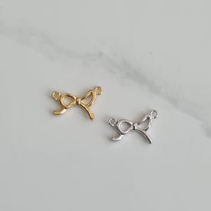 Op de afbeelding: Twee metalen strikbedels, een gouden en een zilveren, met een eenvoudig ontwerp. Deze bedels zijn perfect om een ​​toets van elegantie toe te voegen aan sieradenprojecten.
