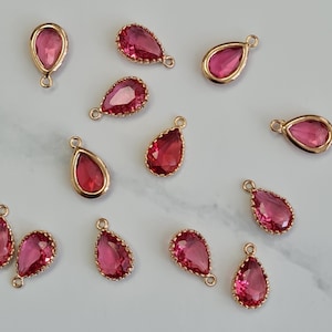 May include: Teardrop-shaped charms with a gold-coloured metal frame and a vibrant pink gemstone centre. The charms have a small loop at the top for attachment to jewellery. The gemstones have a faceted cut, enhancing their sparkle and are suitable for crafting.