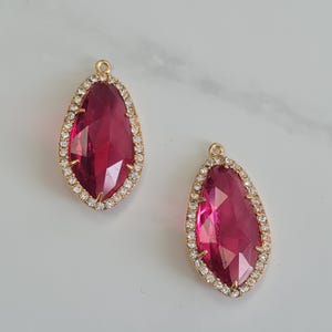 Pode incluir: Dois pingentes em forma de gota com centros facetados rosa escuro. Cada pingente é contornado com pequenos cristais transparentes em metal dourado. Há um pequeno laço no topo de cada pingente.