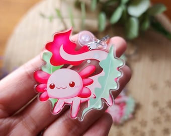 Può includere: Un ciondolo axolotl rosa e verde con uno sfondo di foglie. L'axolotl è un'illustrazione in stile cartone animato con un'espressione carina. Il ciondolo è fatto di acrilico trasparente.