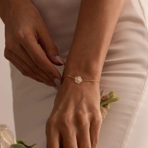 Peut inclure: Un bracelet chaîne en or avec un charm en forme de fleur blanche. Le bracelet est porté au poignet, avec une robe blanche en arrière-plan. Un bijou simple et élégant.