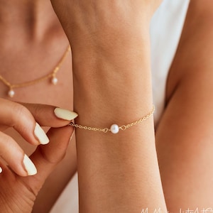 Könnte beinhalten: Ein zartes Armband aus goldener Kette mit einem einzigen weißen Perlenanhänger. Das Armband wird am Handgelenk einer Person gezeigt.