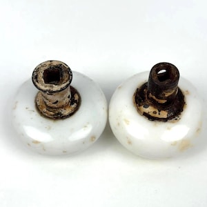 May include: Two round white porcelain door knobs with weathered, dark metallic hardware attachments. The smooth, glossy white surfaces show minor signs of age and discoloration, while the central metal stems feature a square opening.