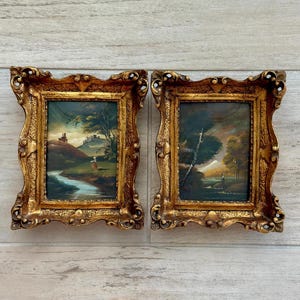 May include: Two rectangular landscape paintings housed in ornate, textured gold frames. The left painting depicts a river flowing through a hilly landscape with a distant structure. The right painting shows a figure in a wooded area with a bright sky.