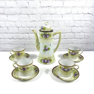 May include: A porcelain tea set consisting of a tall teapot with a lid and four matching teacups with saucers. Each piece features a pale yellow base with hand-painted purple violet flowers, green leaves, and delicate gold trim along the edges.