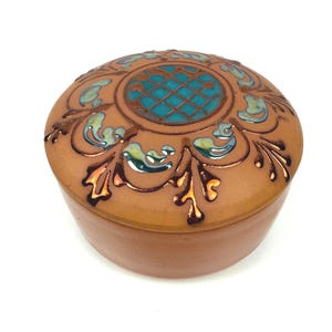 May include: A round terracotta ceramic lidded box featuring a central turquoise grid pattern on the lid, surrounded by embossed floral-like motifs with metallic bronze and blue glaze accents on a smooth, matte tan clay surface.