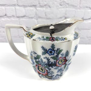 May include: A white ceramic creamer with a faceted body and a gold-trimmed rim. The exterior features a colorful floral pattern with blue, pink, yellow, and green blossoms. A dark, circular lid with a small white knob sits atop the creamer.