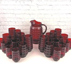 May include: A large ruby red glass pitcher with a textured hobnail pattern and a matching set of twenty-four cylindrical drinking glasses. Each piece features a repeating circular indentation design across the exterior surface of the deep red glass.