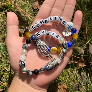 May include: Two beaded bracelets with the names "CORALINE" and "WYBIE" spelled out in white letter beads. The bracelets feature a variety of beads in colors like black, white, blue, and yellow, with a silver skeleton hand charm.