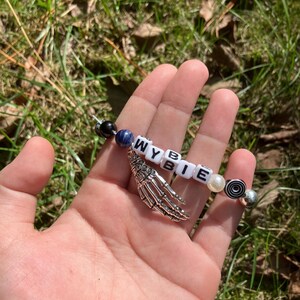 May include: A beaded bracelet with the letters "WYBIE" spelled out in white letter beads. The bracelet includes a silver skeleton hand charm, black, blue, and pearl-colored beads, and a decorative bead with a black and white spiral design.