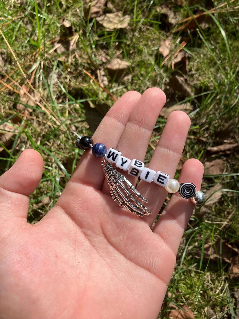 May include: A beaded bracelet with the letters "WYBIE" spelled out in white letter beads. The bracelet includes a silver skeleton hand charm, black, blue, and pearl-colored beads, and a decorative bead with a black and white spiral design.