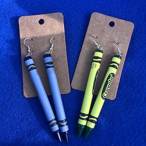 May include: Two pairs of crayon earrings. One pair is blue and the other is yellow and green. The earrings are on brown card stock with the word "Crayola" printed on one of the cards.