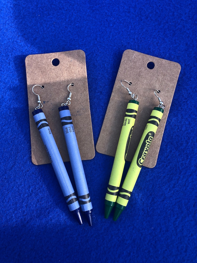 May include: Two pairs of crayon earrings. One pair is blue and the other is yellow and green. The earrings are on brown card stock with the word "Crayola" printed on one of the cards.