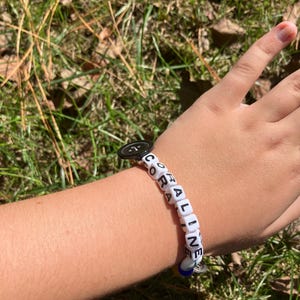 Puede incluir: Una pulsera de cuentas blancas que deletrea "CORALINE" con cubos de letras negras. La pulsera tiene un cierre de bot&oacute;n negro y un peque&ntilde;o dije azul y plateado. La pulsera est&aacute; en una mu&ntilde;eca sobre un fondo de hierba verde.