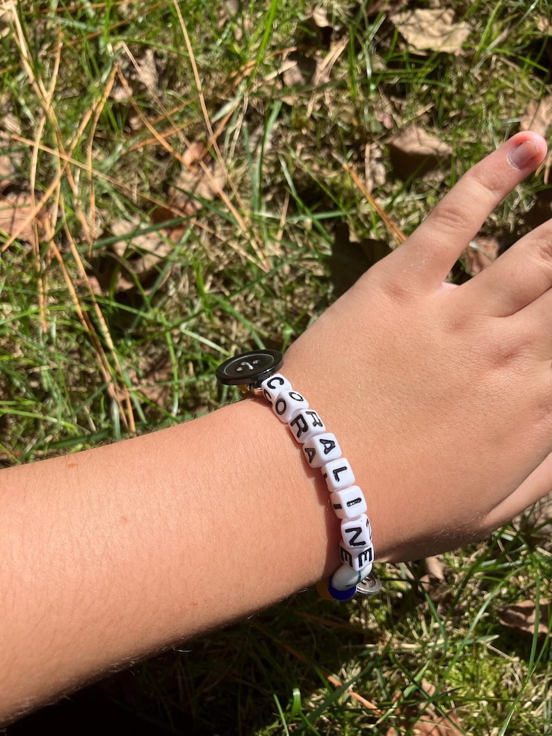 Puede incluir: Una pulsera de cuentas blancas que deletrea "CORALINE" con cubos de letras negras. La pulsera tiene un cierre de bot&oacute;n negro y un peque&ntilde;o dije azul y plateado. La pulsera est&aacute; en una mu&ntilde;eca sobre un fondo de hierba verde.