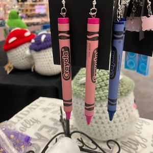 May include: A pair of pink Crayola crayon earrings and a blue Crayola crayon earring. The earrings are hanging from a display stand. The pink crayons are labeled "Rose Delight" and the blue crayon is labeled "Crayola.com".