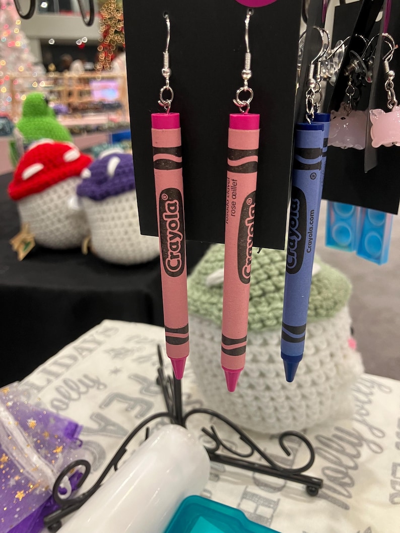 May include: A pair of pink Crayola crayon earrings and a blue Crayola crayon earring. The earrings are hanging from a display stand. The pink crayons are labeled "Rose Delight" and the blue crayon is labeled "Crayola.com".
