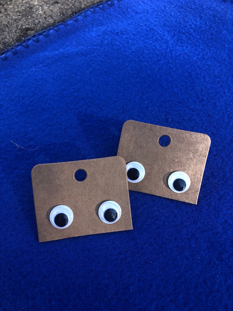 May include: Two pairs of googly eye earrings on brown card stock. The eyes are white with black pupils.