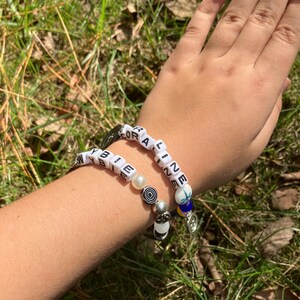 May include: Two beaded bracelets with white letter beads spelling out "CORALINE" and "BIEBIE". The bracelets feature various beads, including pearls and decorative charms. The bracelets are worn on a person's wrist.