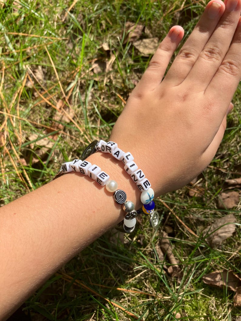 May include: Two beaded bracelets with white letter beads spelling out "CORALINE" and "BIEBIE". The bracelets feature various beads, including pearls and decorative charms. The bracelets are worn on a person's wrist.