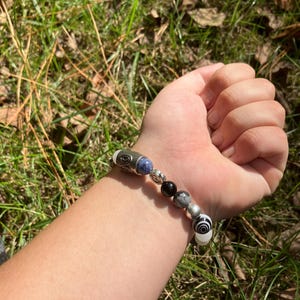 May include: A beaded bracelet featuring a mix of silver, black, white, and blue beads. The bracelet includes a variety of shapes and patterns, including a cylindrical bead with a swirl design. The bracelet is shown on a wrist.