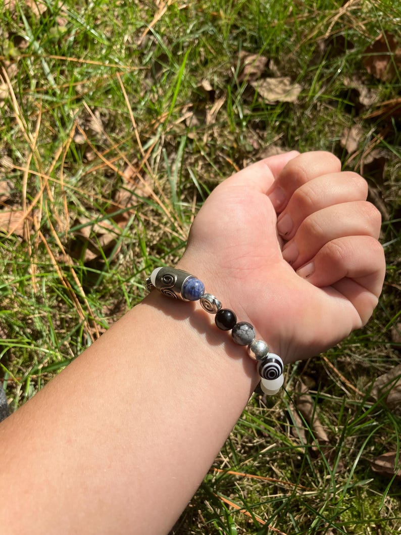 May include: A beaded bracelet featuring a mix of silver, black, white, and blue beads. The bracelet includes a variety of shapes and patterns, including a cylindrical bead with a swirl design. The bracelet is shown on a wrist.